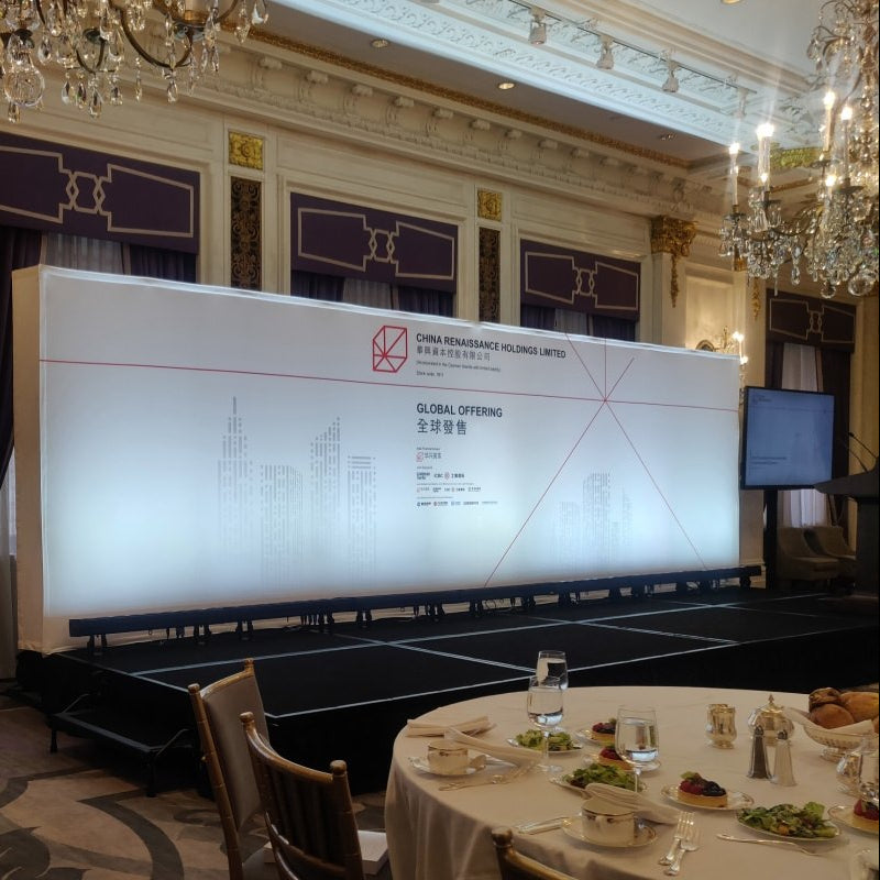 A large white event backdrop screen with red and black text and graphics, set in a decorated banquet hall with crystal chandeliers, dining tables, and digital display screens.