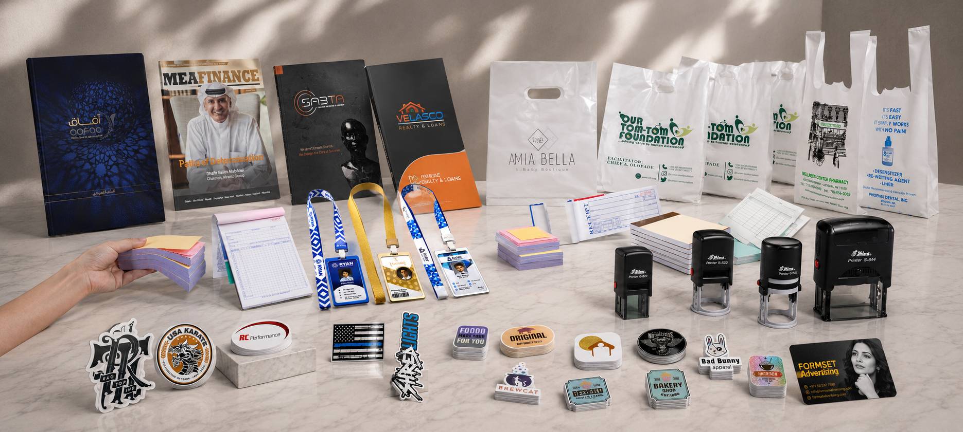 A collection of customized branding and stationery items arranged on a light-colored surface, including printed brochures, branded shopping bags, lanyards, sticky notes, pens, stickers, and self-inking stamps. A hand is visible holding a small notepad on the left side.