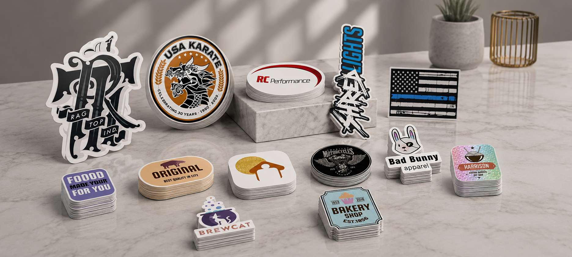 A styled tabletop display of multiple small printed stickers, patches, and coaster-like square items arranged on a light marble surface. The products include novelty designs such as a black-and-white monogram sticker, a circular USA Kamik patch, an RC Performance logo piece, a blue-and-white striped flag patch, bunny-themed stickers, and several bakery/cafe themed square prints.