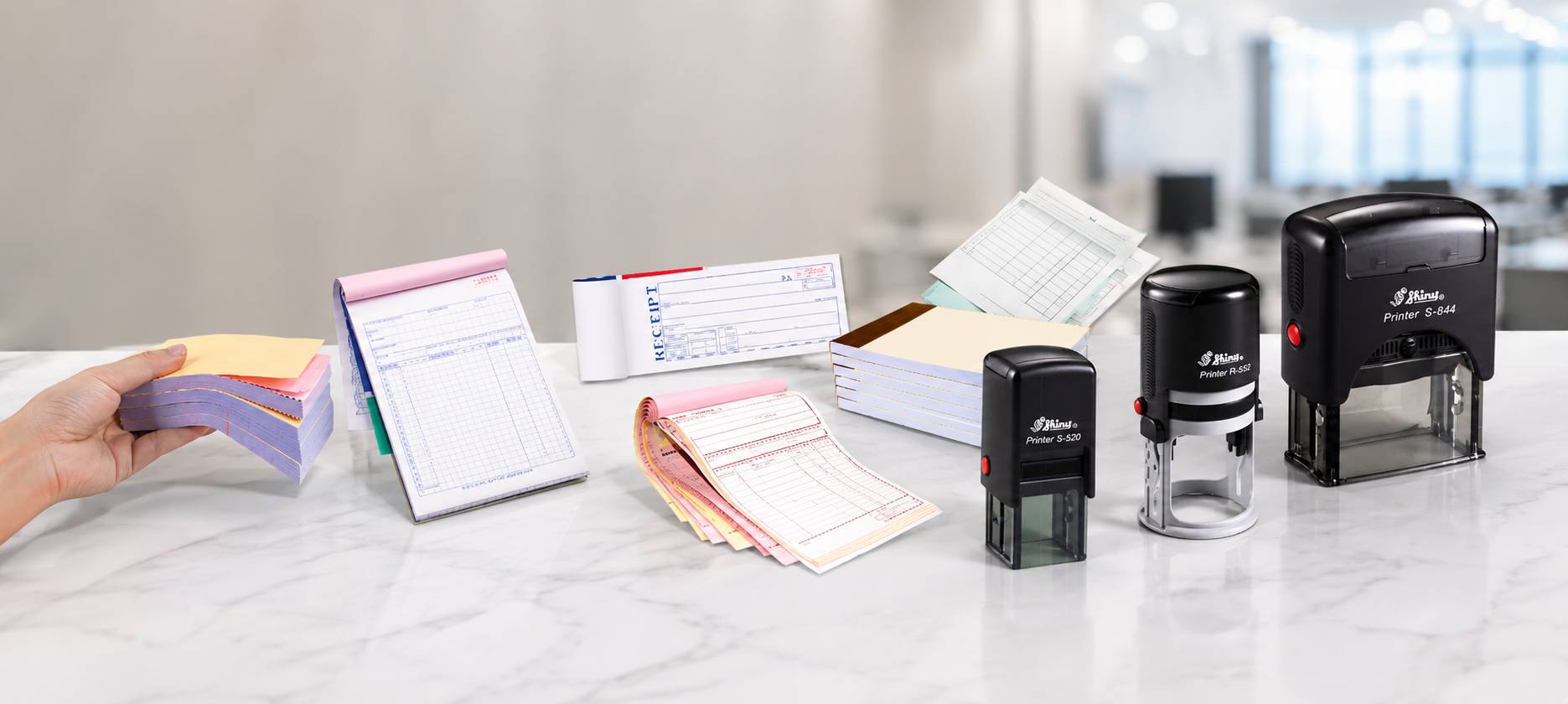 A set of self-inking stamps and notepad stacks displayed on a marble countertop in a bright office setting. A hand is visible holding a stack of pastel notepads on the left, while three black self-inking stamps of different sizes stand on the counter.