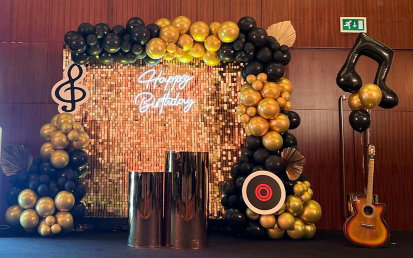 Gold shimmer sequin backdrop with black and gold balloons, LED neon Happy Birthday sign, and musical decor props by Formset Advertising.